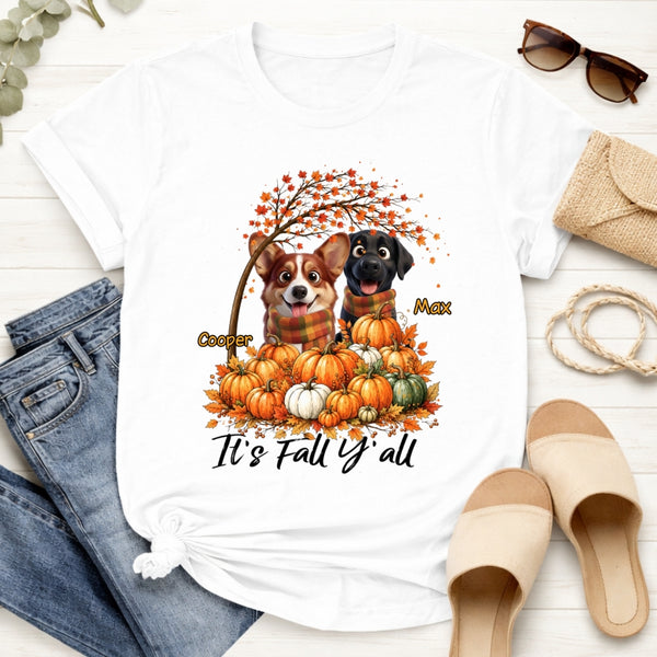 A white graphic T-shirt laid flat on a light wood surface. The shirt features a fall-themed illustration of two dogs among pumpkins with the text 'It's Fall Y'all.