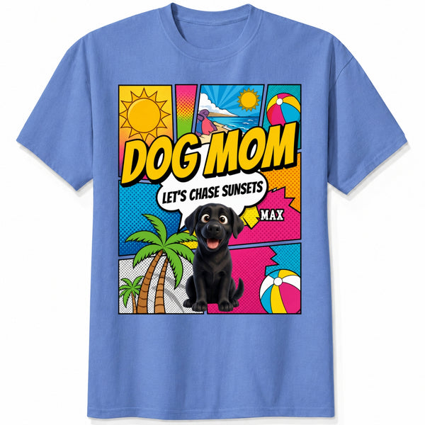 A blue short-sleeve t-shirt featuring a colorful comic-style graphic on the front with the text 'DOG MOM LET'S CHASE SUNSETS' and custom dogs art and dog's names with palm trees, a beach ball, and a sun.