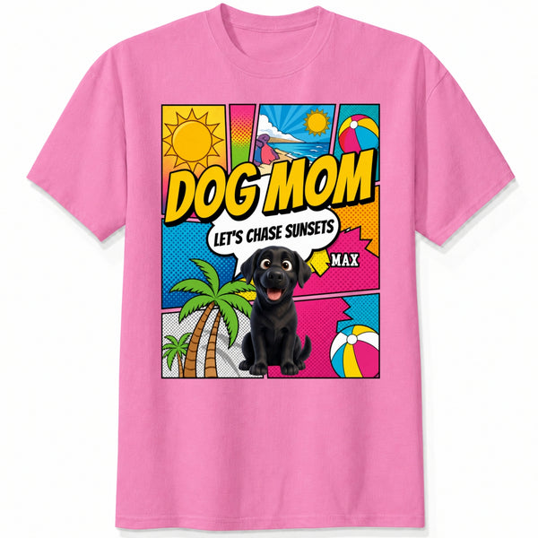 A pink short-sleeve t-shirt featuring a colorful comic-style graphic on the front with the text 'DOG MOM LET'S CHASE SUNSETS' and custom dogs art and dog's names with palm trees, a beach ball, and a sun.