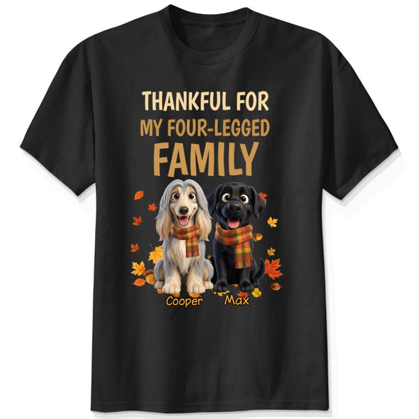 A black short-sleeve crewneck t-shirt with a graphic print. The print features the text 'THANKFUL FOR MY FOUR-LEGGED FAMILY' in beige and orange lettering, above an illustration of custom dogs wearing orange plaid scarves, surrounded by autumn leaves. customized with dog's names