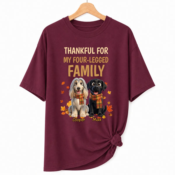 A maroon short-sleeve t-shirt hanging on a wooden hanger against a white background. The shirt features a graphic print with the text 'THANKFUL FOR MY FOUR-LEGGED FAMILY' in gold, above dogs wearing orange plaid scarves, surrounded by small orange and yellow autumn leaves.