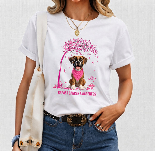 A woman wearing a white graphic T-shirt featuring a dog under a pink tree with the words 'Breast Cancer Awareness.' The photo is cropped from the shoulders to the hips, with blue jeans, a cream tote bag, and gold jewelry visible against a light neutral background.